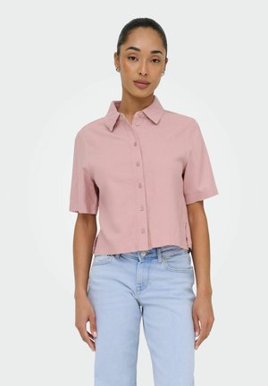 Kobieta w jasnoróżowej koszuli na krótkie rękawy zapinanej na guziki oraz jasnoniebieskich dżinsach, stojąca na tle jednolitego białego tła.