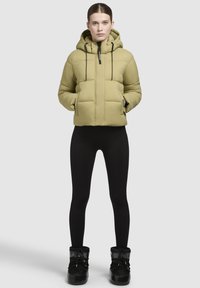 Jachetă căptușită de culoare verde deschis, cu glugă, fermoar frontal și șnururi negre. Pur tată peste colanți negri și cizme negre căptușite cu blană.