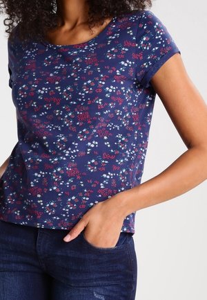 Femme portant un haut à manches courtes bleu marine orné de petits motifs floraux rouges et blancs, associé à un jean bleu foncé, main dans la poche.