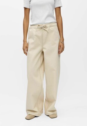 Personne portant un pantalon beige à jambes larges avec une taille à cordon et un haut blanc à manches courtes, debout devant un fond uni.