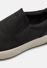 Chaussure noire à enfiler en cuir lisse avec semelle de couleur claire. Présente un design minimaliste, des coutures discrètes et des accents perforés.
