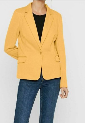 Persoon die een mosterdgele blazer draagt over een zwart topje en blauwe spijkerbroek, staand met één hand achter de rug.