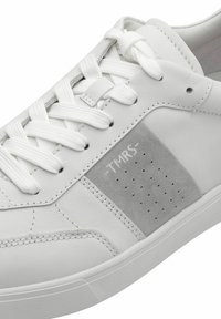 Bílé kožené tenisky s texturovanou šedou semišovou panelem, perforovanými detaily a plochými bílými tkaničkami. Mají gumovou podrážku a prošívané akcenty.