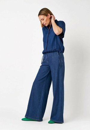 Frau mit blondem Haar trägt ein locker sitzendes dunkelblaues Jeanshemd und weite Jeans, steht und berührt ihr Haar, grüne Schuhe sind sichtbar.