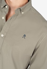 Camisa de botón color caqui claro fabricada con un tejido texturizado que presenta un pequeño logo bordado en color navy, exhibido prominentemente cerca del área del pecho.