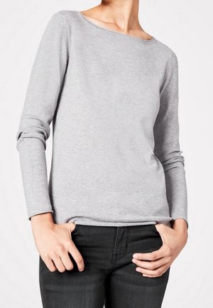 Personne portant un haut gris clair à manches longues et un jean noir, debout les mains dans les poches devant un fond uni clair.