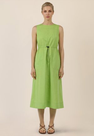 Frau steht mit dem Gesicht nach vorne, trägt ein ärmelloses, hellgrünes Midikleid mit schwarzem Kordelzug in der Taille und braune flache Sandalen.