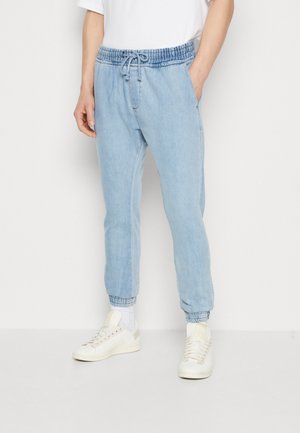 Persona con pantalones jogger de denim azul claro con puños y cintura elásticos, combinados con zapatillas blancas y una camisa blanca.