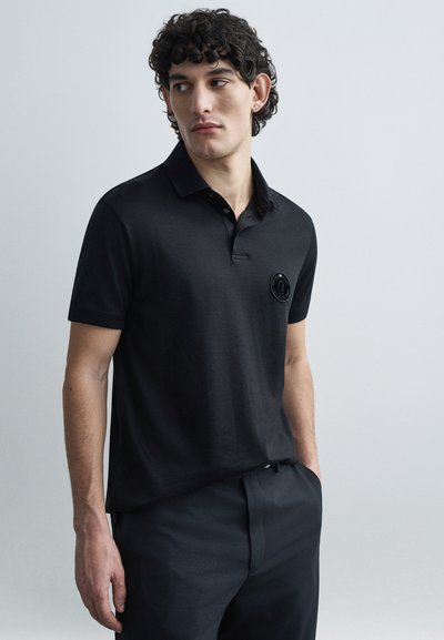 Zwart poloshirt met een korte kraag, voorzien van een ronde logopatch op de linkerbovenkant van de borst. Gemaakt van soepel stof, gecombineerd met een donkere broek.