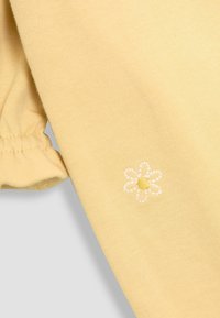Tela amarilla con textura suave, que presenta un puño con volantes y un pequeño diseño de flor bordada en blanco con un centro amarillo.