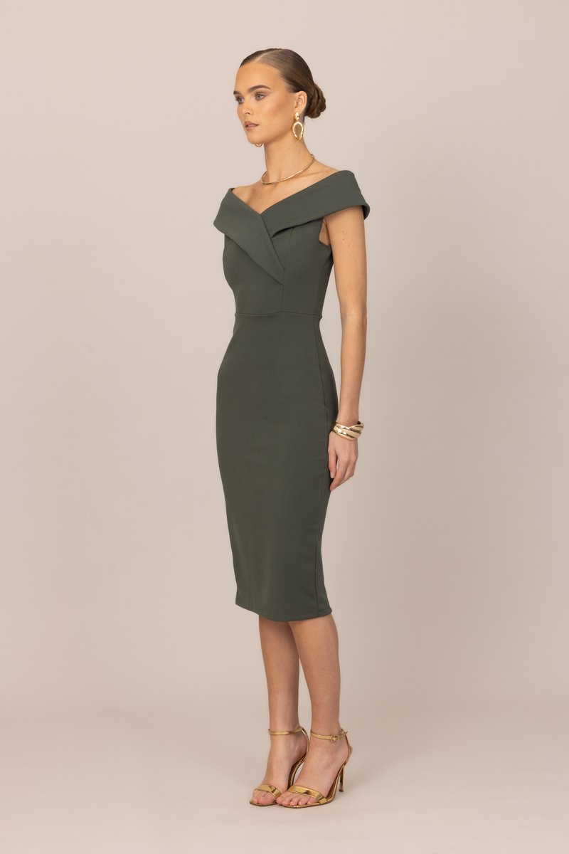 Γυναίκα με μαλλιά σε κότσο φορόντας μεσοφόρι φόρεμα ελιάς με ακάλυπτους τους ώμους, χρυσά σκουλαρίκια, βραχιόλια, κολιέ και σανδάλια με ψηλά τακούνια.