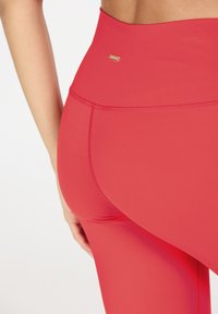 Hoge rode leggins van gladde, rekbare stof met een gouden logo-accent op de tailleband en platte naden voor comfort.