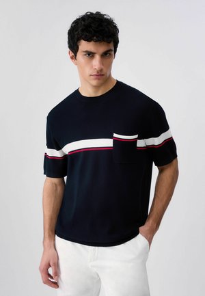 Młody mężczyzna w czarnej koszulce z krótkim rękawem, z białymi i czerwonymi poziomymi paskami oraz pasującą kieszonką, w połączeniu z białymi spodniami.