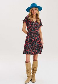 Květinové šaty s černým pozadím a červenými a modrými květy, krátkými volánkovými rukávy, páskem na zavázání v pase a béžovými botami do půli lýtek.