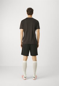 Camiseta atlética preta de mangas curtas, combinada com calções pretos e meias brancas até ao joelho. Chuteiras brancas e laranja com o logo visível.