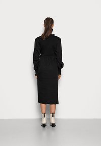 Vestido preto com corpete texturizado, mangas longas bufantes e cintura amarrada. Com um comprimento até o tornozelo e botas de salto bloco em creme.