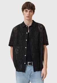 Camisa negra de manga corta con un patrón de encaje que presenta símbolos y letras. Botones en la parte frontal, llevada sobre una camiseta negra, con pantalones de mezclilla claros.