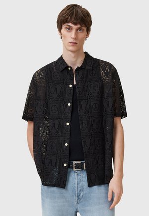 Zwarte T-shirt met korte mouwen en een kantpatroon met symbolen en letters. Knopen aan de voorkant, gedragen over een zwarte top, met lichte spijkerbroek.
