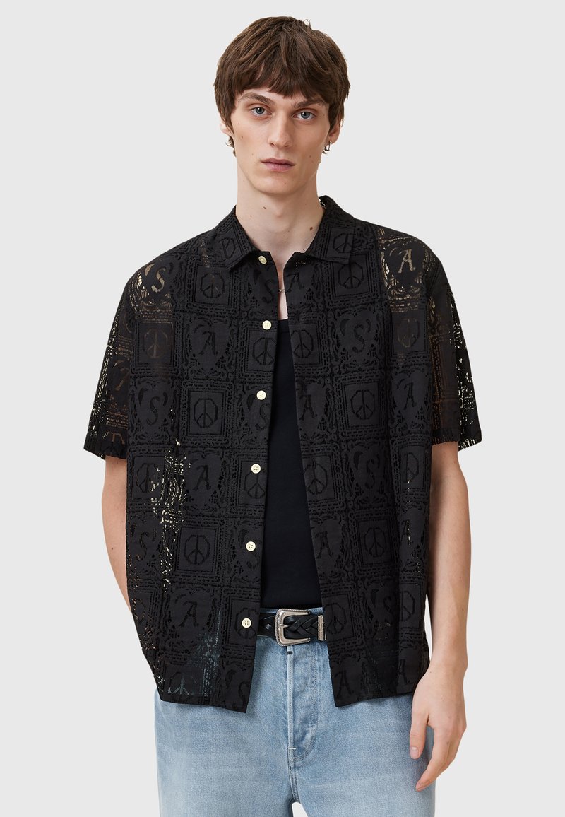 Camisa negra de manga corta con un patrón de encaje que presenta símbolos y letras. Botones en la parte frontal, llevada sobre una camiseta negra, con pantalones de mezclilla claros.