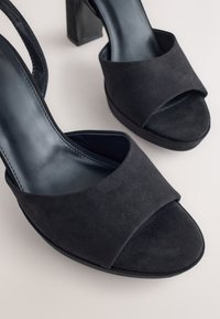 Sorte åbne slingback-hæle i ruskind med en tyk blokhæl og bred forrem, med en glat inderforing og subtil tekstur.