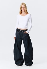 Maglietta bianca a maniche lunghe con scollo a V; pantaloni larghi in denim blu scuro oversize; cintura nera con fibbia in argento; modello in piedi con le mani nelle tasche.