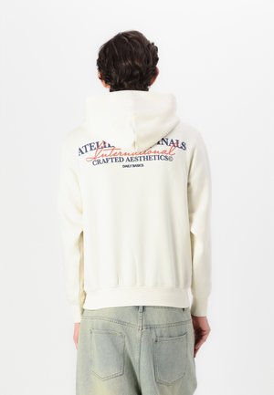 Haut à capuche couleur crème avec un cordon de serrage, affichant un texte multicolore dans le dos. Tissu doux, poignets et ourlet côtelés ; coupe décontractée.