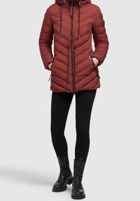 Chaqueta acolchada roja con capucha, cordones ajustables, bolsillos con cremallera, detalles en negro, y combinada con leggings negros y botas de tobillo.