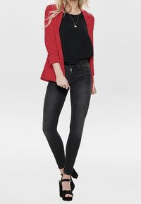 Blazer rojo sobre un top negro, combinado con jeans skinny negros y sandalias de tacón alto negras. Un collar simple acentúa el conjunto.