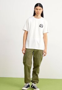 Camiseta blanca de algodón con un logo gráfico negro en el lado izquierdo del pecho, combinada con pantalones cargo verde olivo que cuentan con múltiples bolsillos.
