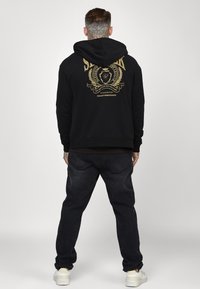 Sudadera negra con un gráfico dorado en la parte trasera, acompañada de jeans oscuros y zapatillas blancas, vista desde la parte de atrás.
