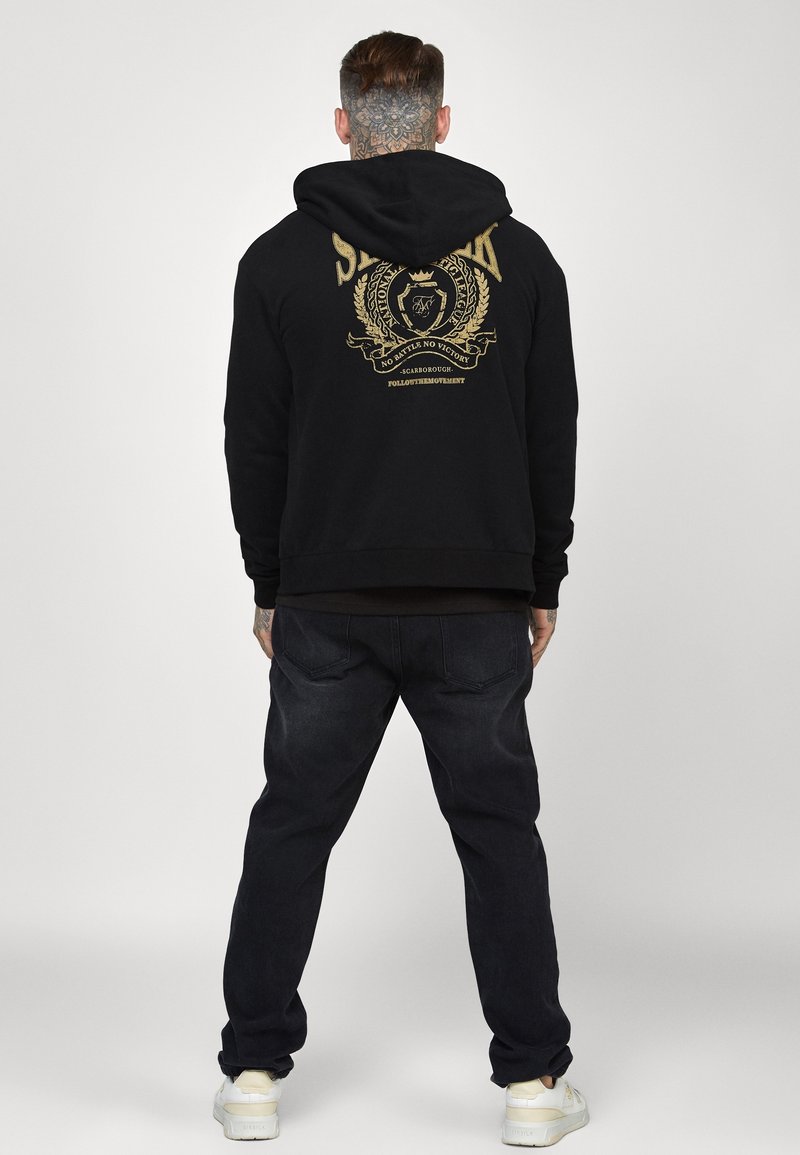 Sudadera negra con un gráfico dorado en la parte trasera, acompañada de jeans oscuros y zapatillas blancas, vista desde la parte de atrás.