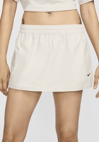 Egy nő fehér, elasztikus derékú teniszsprüttben áll, a bal oldalán egy kicsi fekete Nike logóval, a köldöke látható, amelyen piercing van.