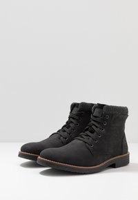 Bottines noires avec une tige texturée, bout rond, fermeture à lacets à l'avant et un doux revers gris. Semelle en caoutchouc avec un accent brun clair.
