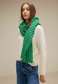 Sciarpa verde a scacchi con dettagli grigi, avvolta attorno al collo, abbinata a un maglione crema e jeans blu.