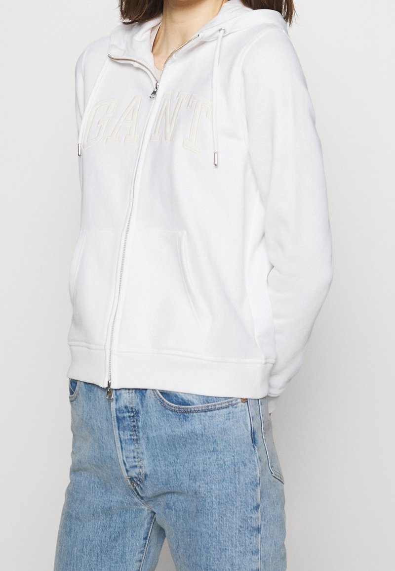White zip-up hoodie with drawstrings, featuring a textured "GANT" logo in beige. Soft fabric and front pockets, paired with light blue jeans.