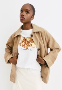 Lichtbruine suède jas met een kraag en twee knoopsluitingen, gedragen over een wit T-shirt met een toastafbeelding en tekst. Glad oppervlak.