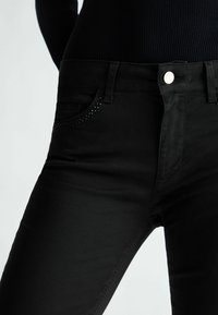 Primer plano de pantalones ajustados negros con pequeñas cuentas negras decorando el borde del bolsillo frontal, llevados con una blusa negra de canalé.