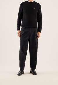 Maglione nero a maglia con scollatura tonda e un piccolo emblema dorato, abbinato a pantaloni neri a vestibilità ampia e scarpe in pelle nere.