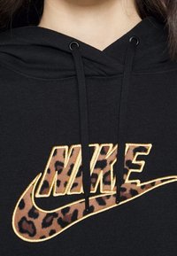 Sudadera negra con un logo de Nike bordado en dorado y estampado de leopardo, con cordones negros. Textura suave y diseño casual.