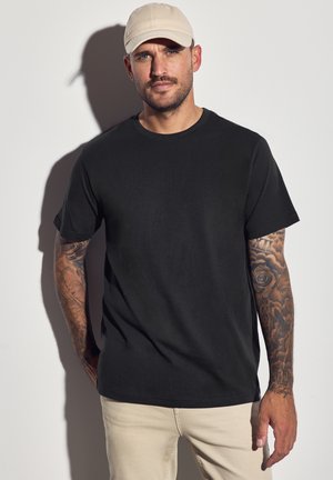 Zwarte katoen T-shirt met korte mouwen, ronde halslijn, gecombineerd met licht beige broek. Opvallende tatoeage detail op de rechterarm, minimalistisch ontwerp.