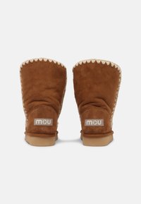 Bottes ankle en daim marron avec des coutures crème et doublure en laine de mouton. Extérieur texturé et semelle en caoutchouc plate de couleur tan. Patch logo à l'arrière.