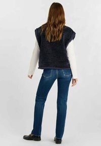 Donkerblauwe gebreide vest met een losse pasvorm, gedragen over een witte langoeshirt, gecombineerd met donkerblauwe spijkerbroek en zwarte instapschoenen. Golvend haar zichtbaar.