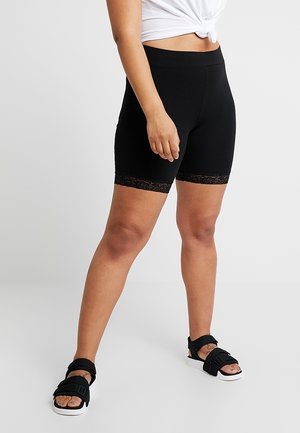 Person iført sorte shorts til midt på låret med blonder, hvid bundet crop top og sorte åbne sandaler, stående mod en ensfarvet baggrund.
