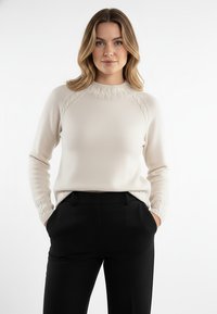 Kremowy sweter z dzianiny z wysokim kołnierzem i teksturowanymi akcentami na ramionach i mankietach, zestawiony z czarnymi, dopasowanymi spodniami.