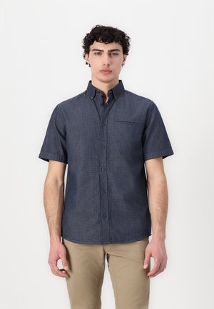 Chemise à manches courtes en tissu bleu foncé avec un motif texturé, fermée par des boutons à l'avant et dotée d'une seule poche poitrine. Portée avec un pantalon kaki clair.