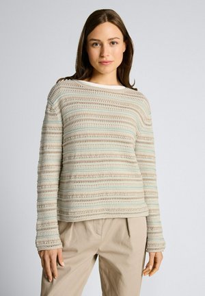 Vrouw staand, gekleed in een gestreepte beige, witte en lichtblauwe gebreide trui en beige broek, vóór een effen lichtgrijze achtergrond.