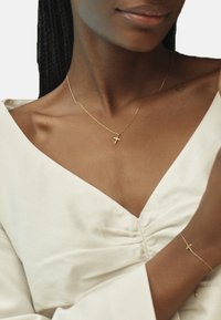 Collana e bracciale con croce d'oro su una persona, caratterizzati da un design di catena delicato. Top bianco con scollatura a V profonda. Texture della pelle liscia.