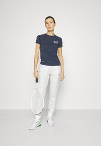 Marineblauwe sportshirt met EA7-logo, gecombineerd met witte met patroon gemaakte broek, terwijl hij een tennisracket vasthoudt en witte sneakers met groene accenten draagt.