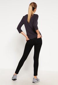 Czarne dopasowane spodnie o gładkiej fakturze, zestawione z długim, szarym topem, noszone z szarymi butami do biegania. Model stoi tyłem.