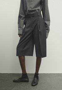 Culottes plissés gris avec une taille élégante, fabriqués à partir d'un tissu texturé. Porté avec des chaussures et des chaussettes noires, sur un fond neutre.